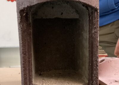 Metal kancalı kahverengi ahşap bir kuş evi, masanın üzerinde duruyor. Açık bir girişi ve alt kısmında şeffaf bir tüpü var.