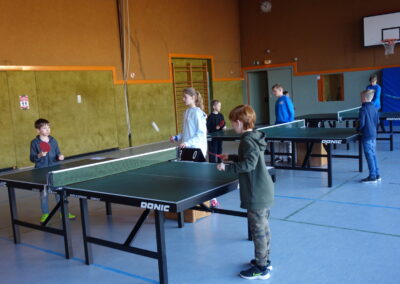 Beş çocuk spor salonunda masa tenisi oynuyor; masalar arasında ping pong topları uçuşurken, onlar da oyuna yoğunlaşıyorlar.
