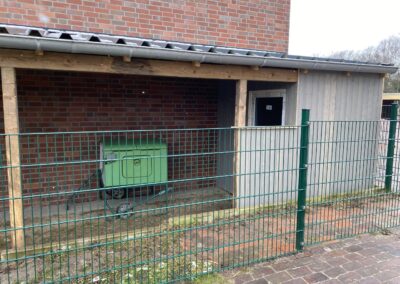 Eine grüne Box steht unter einem überdachten Bereich eines Gebäude an einer Ziegelwand; der Eingang ist mit einem Gitterzaun abgegrenzt.